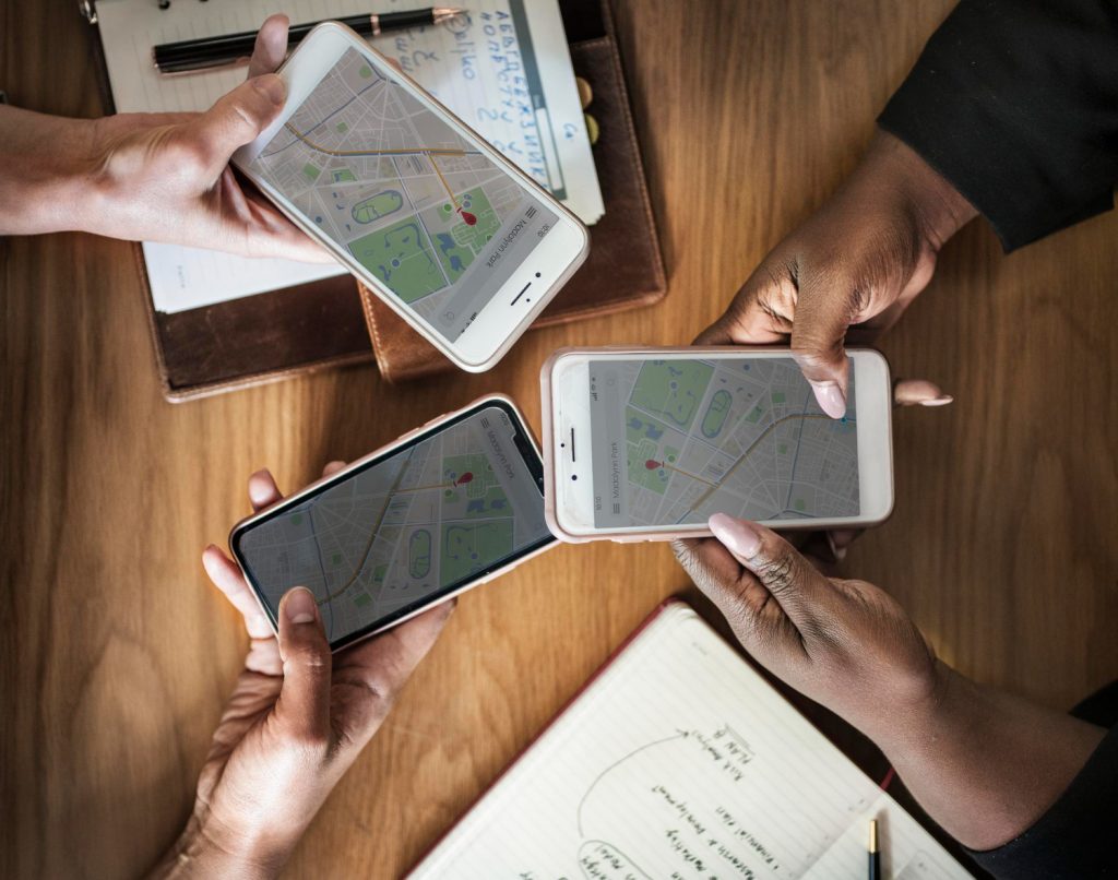 Imagem de uma mesa vista de cima com mãos de três pessoas usando mapas em seus celulares enquanto fazem anotações, ilustrando a função de Programador de Rota.